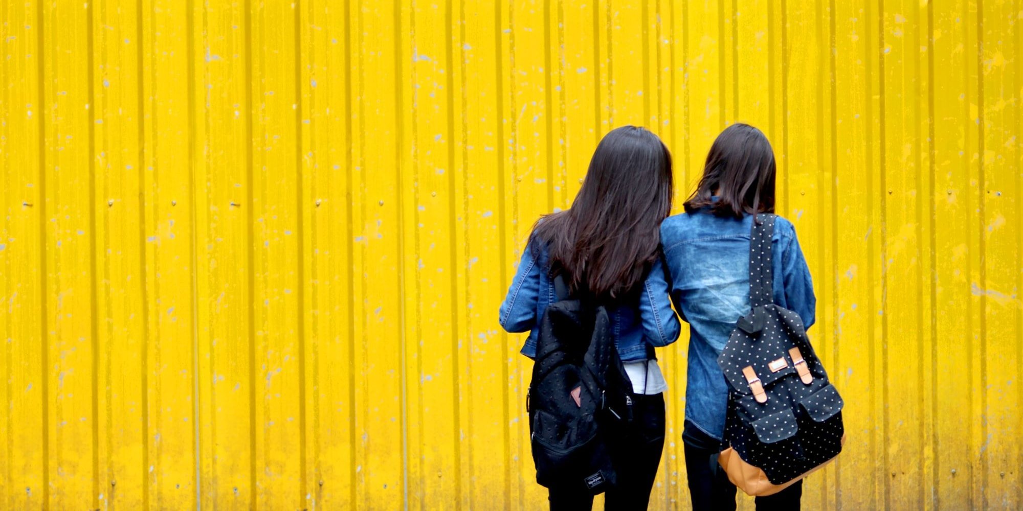 two girls with backpacks standing in front of a yellow wall